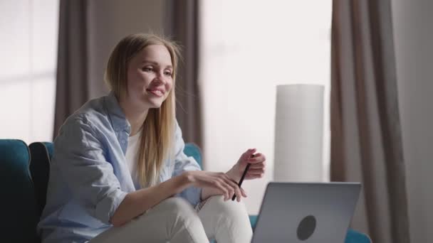 Mujer sentada en un sofá en casa haciendo videollamadas usando un ordenador portátil en el aislamiento personal. Feliz joven rubia profesora mujer trabajando desde casa, hablando en la videoconferencia portátil. — Vídeo de stock