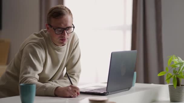 Um homem com óculos trabalha de casa em sua cozinha usando um laptop e a Internet. Trabalho remoto e aprendizagem remota — Vídeo de Stock