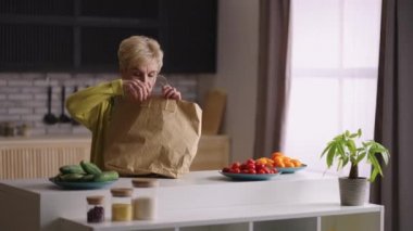 Yaşlı kadın süpermarketten dönüyor ve yiyecek dolu çantalarını açıyor, mutfak masasına eşya koyuyor.