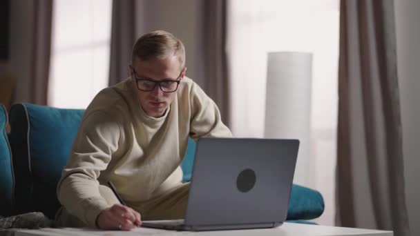 Grave giovane freelance che lavora su freelance da casa digitando e-mail sul computer portatile, uomo concentrato che utilizza il computer per studiare online a casa seduto sul divano, utente maschile occupato sul lavoro a distanza su internet — Video Stock