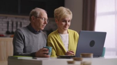 İki emekli ev mutfağında dizüstü bilgisayar kullanıyor, internette sörf yapıyor, internetten alışveriş yapıyor, yaşlı adam ve kadın resimlerini sergiliyor