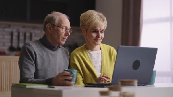 Due pensionati stanno usando laptop a casa cucina, navigazione internet, shopping online, vecchio e donna stanno visualizzando le immagini in mostra — Video Stock