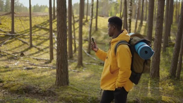 Een zwarte man loopt met de telefoon door het bos en kijkt naar de navigatietoepassing. Afrikaans-Amerikaanse man met een rugzak in slow motion lopen door het bos met een mobiele telefoon — Stockvideo