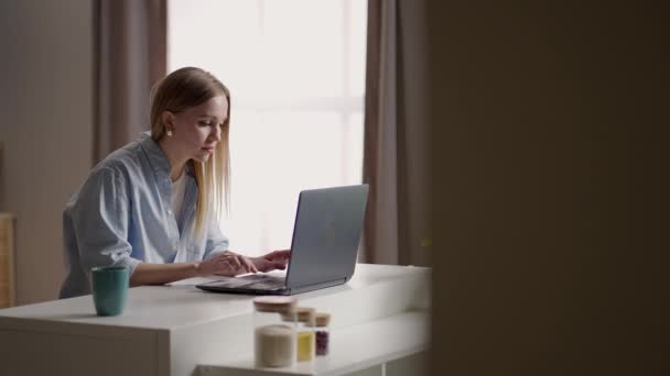 Mobilne biuro w domu. Młoda kobieta siedząca w kuchni w domu, pracująca na laptopie. Styl życia dziewczyna studiuje lub pracuje w pomieszczeniach. Koncepcja kwarantanny prowadzenia działalności na własny rachunek — Wideo stockowe