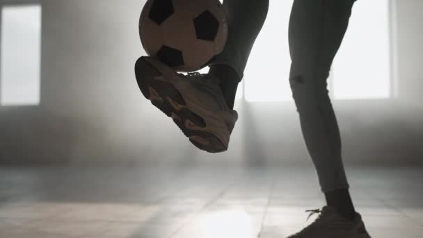 Ein schwarzer Mann in Großaufnahme jongliert in der Garage mit einem Fußball im Sonnenlicht. Fußball-Freestyle bei Sonnenuntergang in einer städtischen Umgebung. Staub und Sonnenuntergang — Stockvideo