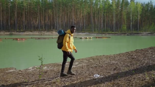 Egy fiatal fekete férfi sárga kurzorban felmászik egy hátizsákkal felfelé az erdőben, és a tavat nézi. Lassú mozgás. — Stock videók