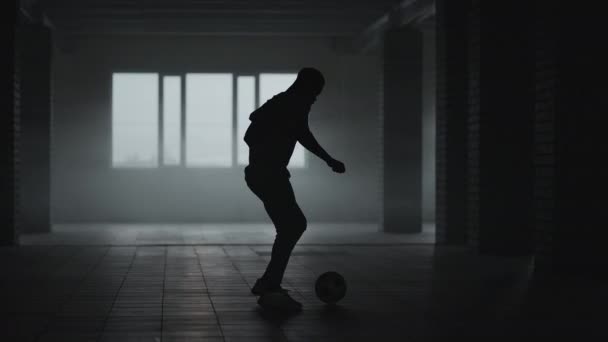 Un footballeur afro-américain s'entraîne à donner des coups de pied et se déplace dans un parking couvert vide. Ralenti homme noir travailler sur le football dribble dans parkin souterrain dans la lumière du soleil. — Video