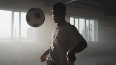 Yakın plan siyahi bir adam garajda güneş ışığında futbol topuyla hokkabazlık yapıyor. Şehir ortamında gün batımında serbest futbol. Toz ve gün batımı