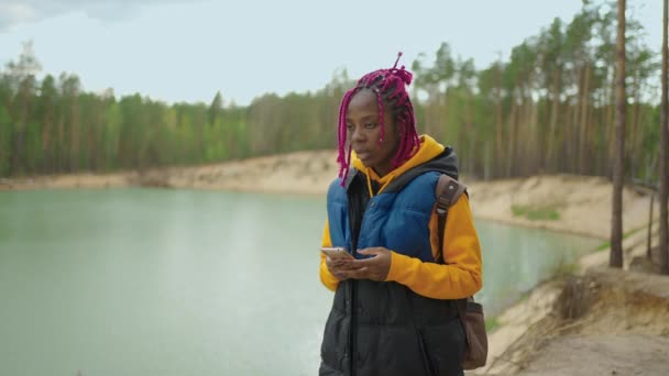 一个年轻的黑人妇女在山中的森林里徒步旅行。用智能手机拍一张山的照片。积极的生活方式。穿着格子衬衫，头戴旅行帽. — 图库视频影像