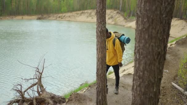 Чёрный мужчина с рюкзаком пользуется мобильным телефоном во время прогулки по лесу через озеро. Фотография и блог — стоковое видео