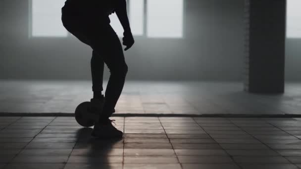 Un footballeur afro-américain s'entraîne à donner des coups de pied et se déplace dans un parking couvert vide. Ralenti homme noir travailler sur le football dribble dans parkin souterrain dans la lumière du soleil. — Video