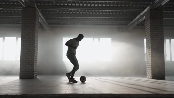 Молодой чёрный мужчина тренируется играть в футбол на подземной парковке под солнечным светом в замедленной съемке. Беги за мячом. — стоковое видео