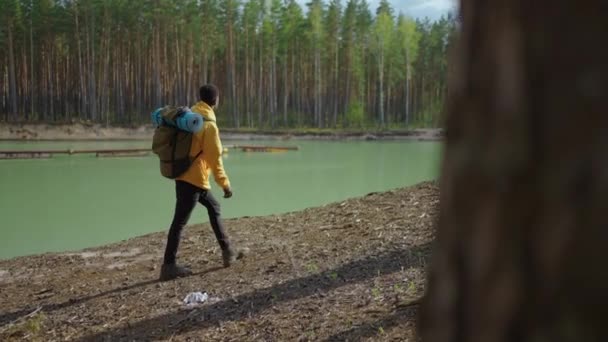 Un joven negro en un cursor amarillo sube con una mochila cuesta arriba en el bosque mirando el lago. Movimiento lento — Vídeo de stock