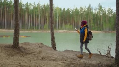 Sırt çantalı siyah bir kız gölün karşısındaki ormanda yürürken cep telefonu kullanıyor. Fotoğraf çek ve blog yaz.