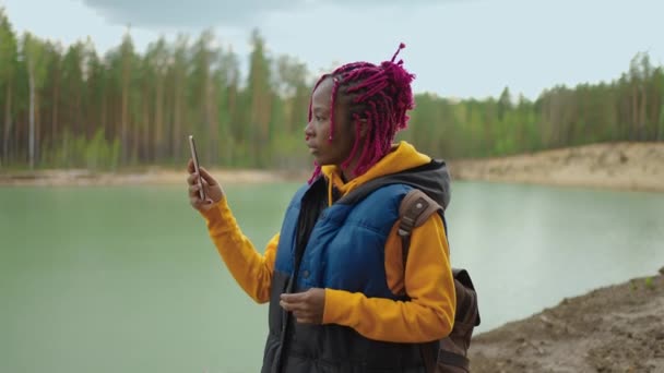 Egy fekete lány hátizsákkal mobiltelefont használ, miközben a tó túloldalán lévő hegyekben túrázik az erdőben. Fotózz és blogolj! — Stock videók