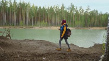 Sarı imleci olan genç siyah bir kadın ormanda tepeye tırmanır ve göle bakar. Yavaş çekim