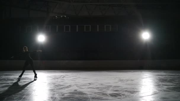 Schlittschuhläufer trainieren im Dunkeln in der Eissporthalle mit Scheinwerfern. Eiskunstlauf im Stadion. Mädchen führt Eiskunstlauf-Elemente auf — Stockvideo