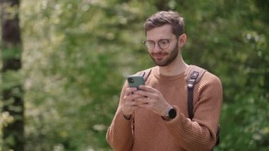 Gözlüklü gülümseyen ve sakallı bir adam sırt çantasıyla ormanda yürüyor ve cep telefonuna yavaş çekimde bir mesaj basıyor..