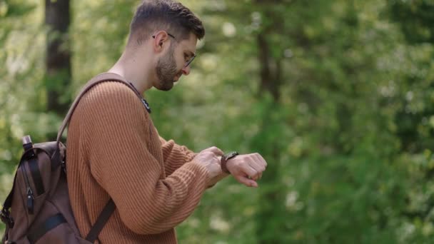 Sırt çantalı ve gözlüklü genç sakallı bir adam, GPS 'i ayarlamak ve ayarlamak için telefonunu kullanarak akıllı bir saati ayarlar. Modern teknoloji saatle yön buluyor. Etkinlik kontrolü — Stok video