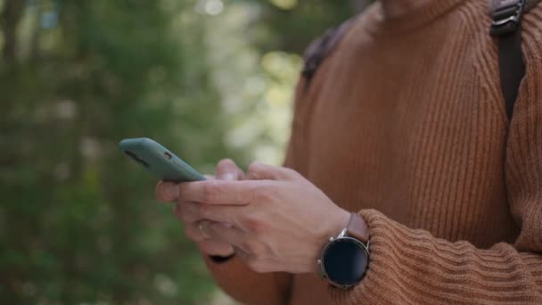 Lächelnder Mann mit Brille und Bart spaziert mit Rucksack durch den Wald und druckt in Zeitlupe eine Nachricht auf sein Handy. — Stockvideo