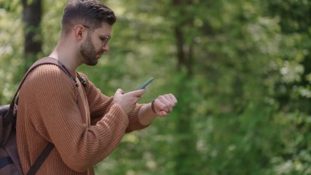 Ein junger bärtiger Mann mit Brille und Rucksack justiert mit seinem Handy eine Smartwatch, um die GPS-Signale zu synchronisieren und anzupassen. Moderne Technik navigiert mit der Uhr. Aktivitätsprüfung — Stockvideo