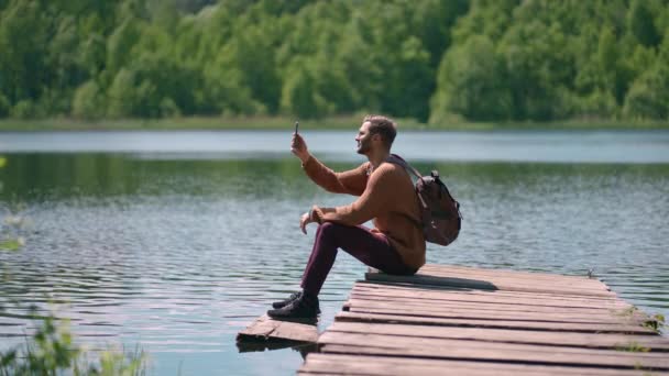 Храбрый человек фотографирует в лесном озере со смартфона, фотографируя живописный пейзаж на фоне природы, наслаждаясь отпускными путешествиями. Медленное движение — стоковое видео