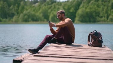 Gözlüklü ve sırt çantalı genç sakallı bir adam ahşap bir göl iskelesinde ağır çekimde oturuyor ve cep telefonuna yol gösteren bir mesaj basıyor.