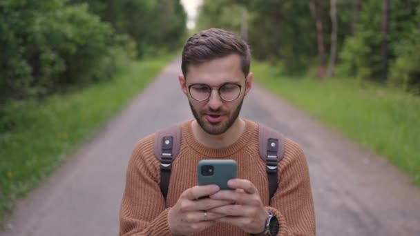 Χαμογελαστός άντρας με γυαλιά με γενειάδα περπατάει στο δάσος με ένα σακίδιο και τυπώνει ένα μήνυμα στο κινητό του σε αργή κίνηση. — Αρχείο Βίντεο