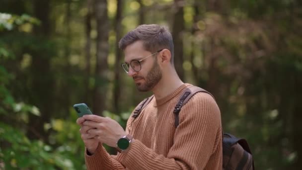 Uśmiechnięty mężczyzna w okularach z brodą spaceruje po lesie z plecakiem i drukuje wiadomość na telefonie komórkowym w zwolnionym tempie. — Wideo stockowe