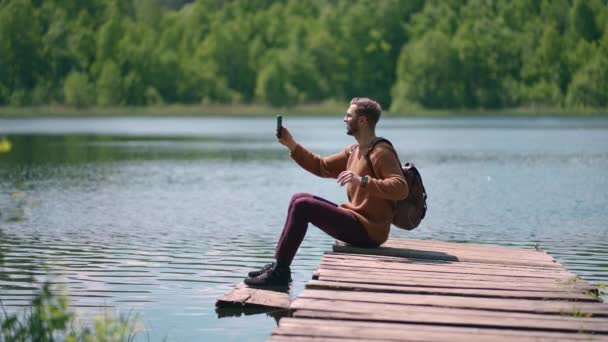 Чоловік з рюкзаком сидить на причалі озера і телефонує за допомогою відеозв'язку на мобільному телефоні. Сидячи на озері в природі, щоб викликати друзів. Блогер знімає вміст каналу — стокове відео
