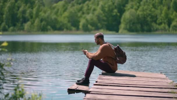 Молодий бородатий чоловік в окулярах і рюкзак сидить на дерев'яному озерному пірсі в повільному русі і друкує повідомлення, що прокладає шлях у мобільному телефоні — стокове відео