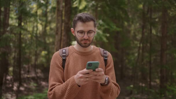 Facet chodzi po lesie i pisze wiadomość przez komórkę. Turysta z plecakiem drukuje wiadomość w telefonie podczas wędrówki. — Wideo stockowe