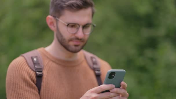 Un uomo cammina nel bosco e scrive un messaggio sul suo cellulare. Turista con uno zaino stampa un messaggio nel suo telefono durante un'escursione — Video Stock