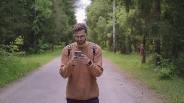 Uśmiechnięty mężczyzna w okularach z brodą spaceruje po lesie z plecakiem i drukuje wiadomość na telefonie komórkowym w zwolnionym tempie. — Wideo stockowe