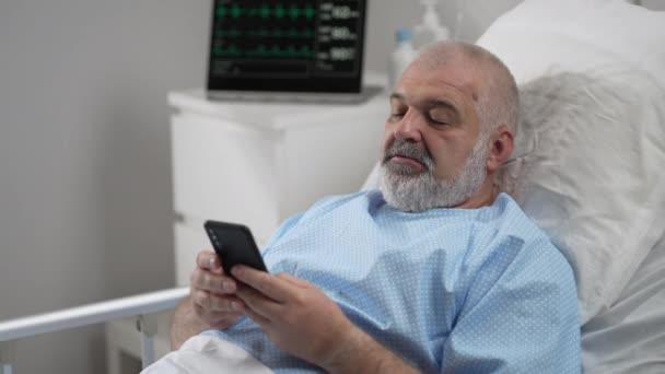 Male Patient Using Mobile Phone In The Ward At Hospital. Senior Male patient holding mobile phone while resting on bed in hospital — ストック動画