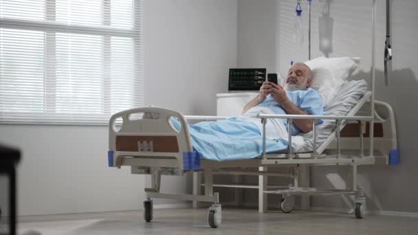Male Patient Using Mobile Phone In The Ward At Hospital. Senior Male patient holding mobile phone while resting on bed in hospital — Stock Video