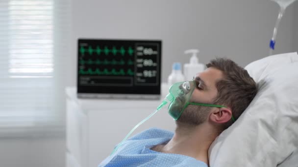 The man lies on the bed in the hospital in oxygen masks and regains consciousness. Connected to the breathing and heart rate monitor lies in the hospital on the bed with his eyes closed — 图库视频影像