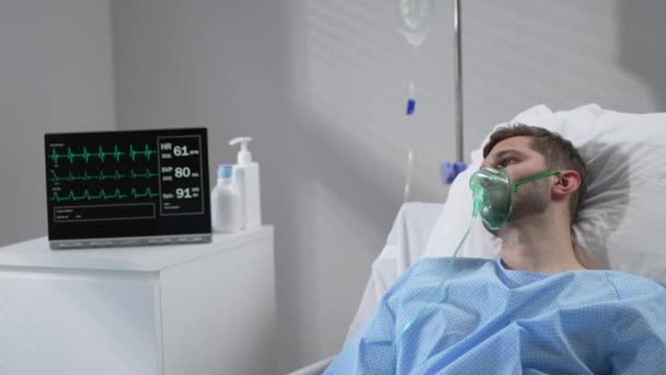 The man lies on the bed in the hospital in oxygen masks and regains consciousness. Connected to the breathing and heart rate monitor lies in the hospital on the bed with his eyes closed — 图库视频影像