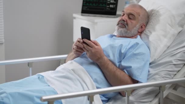 Male Patient Using Mobile Phone In The Ward At Hospital. Senior Male patient holding mobile phone while resting on bed in hospital — Vídeo de Stock