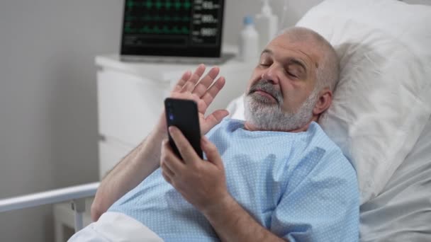 Hospital Ward: senior man Resting in Bed uses Smartphone for Video Call Conference Talk with Family and Friends. video call in hospital room — Video