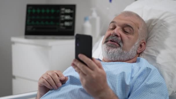 Hospital Ward: senior man Resting in Bed uses Smartphone for Video Call Conference Talk with Family and Friends. video call in hospital room — Stock video