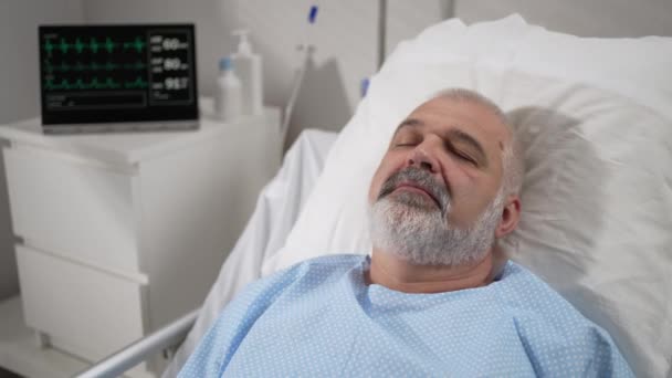 The face of an elderly man lying on a bed in a hospital, unconscious, asleep. The ECG machine shows vital signs — Vídeo de Stock