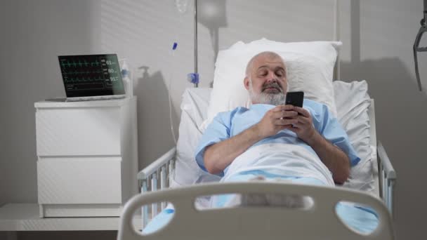 Male Patient Using Mobile Phone In The Ward At Hospital. Senior Male patient holding mobile phone while resting on bed in hospital — Video