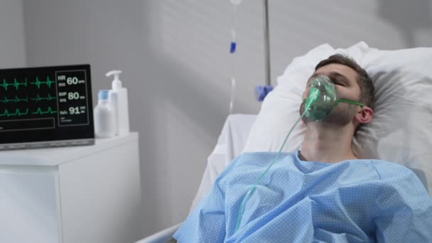 The man lies on the bed in the hospital in oxygen masks and regains consciousness. Connected to the breathing and heart rate monitor lies in the hospital on the bed with his eyes closed — Stok video