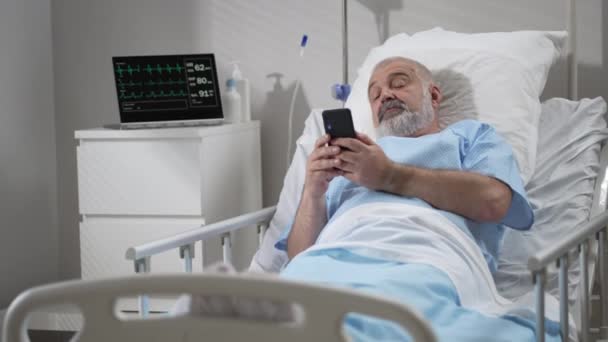 Лікарня Уорд: старший чоловік Відпочинок в ліжку використовує смартфон для відеодзвінків Конференція Спілкування з сім'єю та друзями. відеодзвінок у лікарняній кімнаті — стокове відео