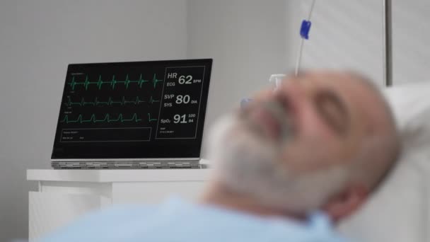 The face of an elderly man lying on a bed in a hospital, unconscious, asleep. The ECG machine shows vital signs — Stockvideo