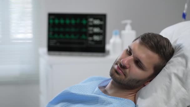 A young man lies on a bloodied hospital and opens his eyes. apparatus for monitoring heart, oxygen and pulse. A young patient of the clinic in a coma. Regain consciousness — Αρχείο Βίντεο