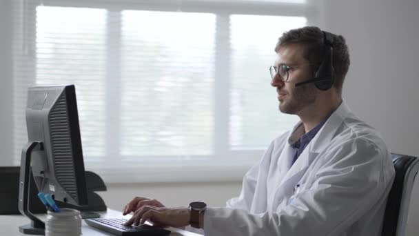 Male doctor or nurse with headset and computer working at hospital. Close up of medical operator with headphone consulting patients. Medical man operator working in a medicine call centre — Stockvideo