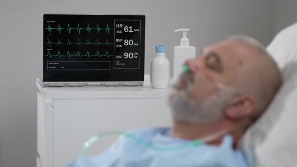 The face of an elderly man lying on a bed in a hospital, unconscious, asleep. The ECG machine shows vital signs — Vídeos de Stock