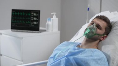 Caucasian man with oxygen mask on lying in bed with white linen, sleeping disturbingly, moving head. Portrait of a man in an oxygen mask who lies on a bed in a hospital.
